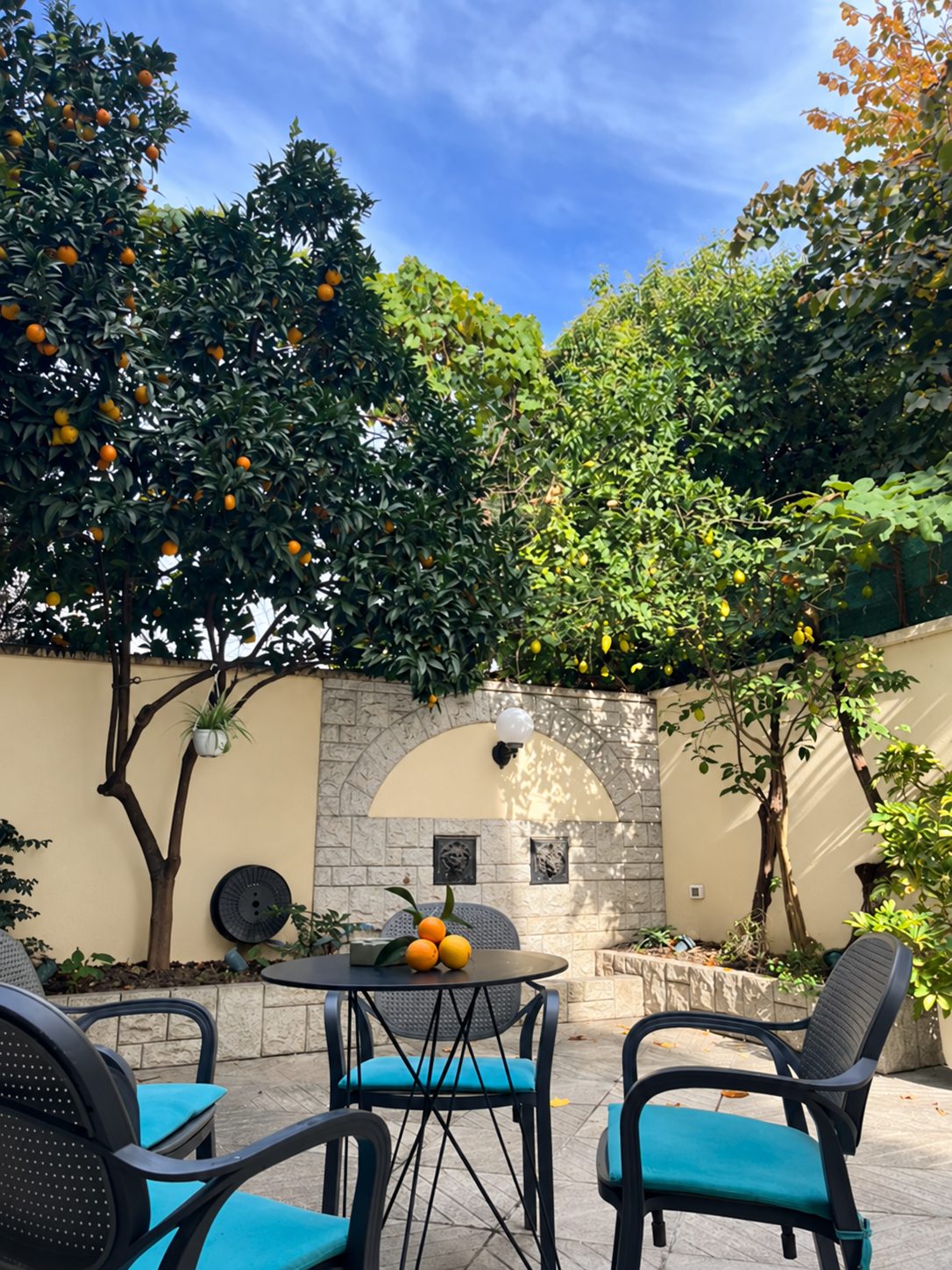 Walled courtyard with citrus trees, a round table with fresh oranges and chairs with turquoise cushions